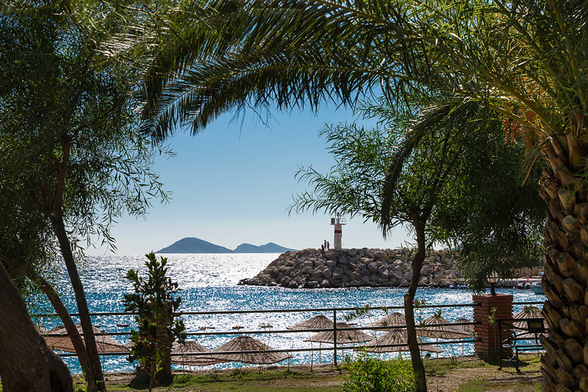 View of gently shelving public beach in Kalkan, Turkey, with turquoise waters, gentle waves, and loungers beneath umbrellas.