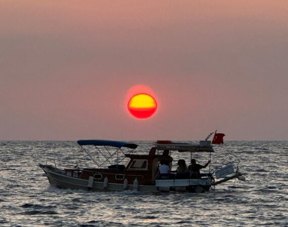 Blood-orange sunset over the sea beyond Kalkan, with a gulet silhouetted near Mouse and Snake Islands against a glowing horizon.