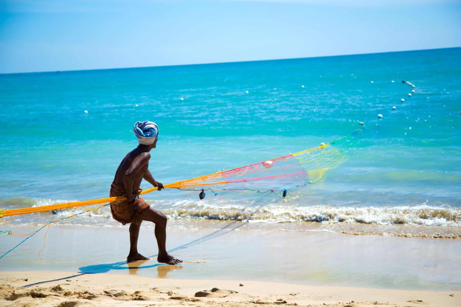Sri Lankan fisherman hauling nets on a white sand beach beneath a bright blue sky, with the azure Indian Ocean stretching out behind him.